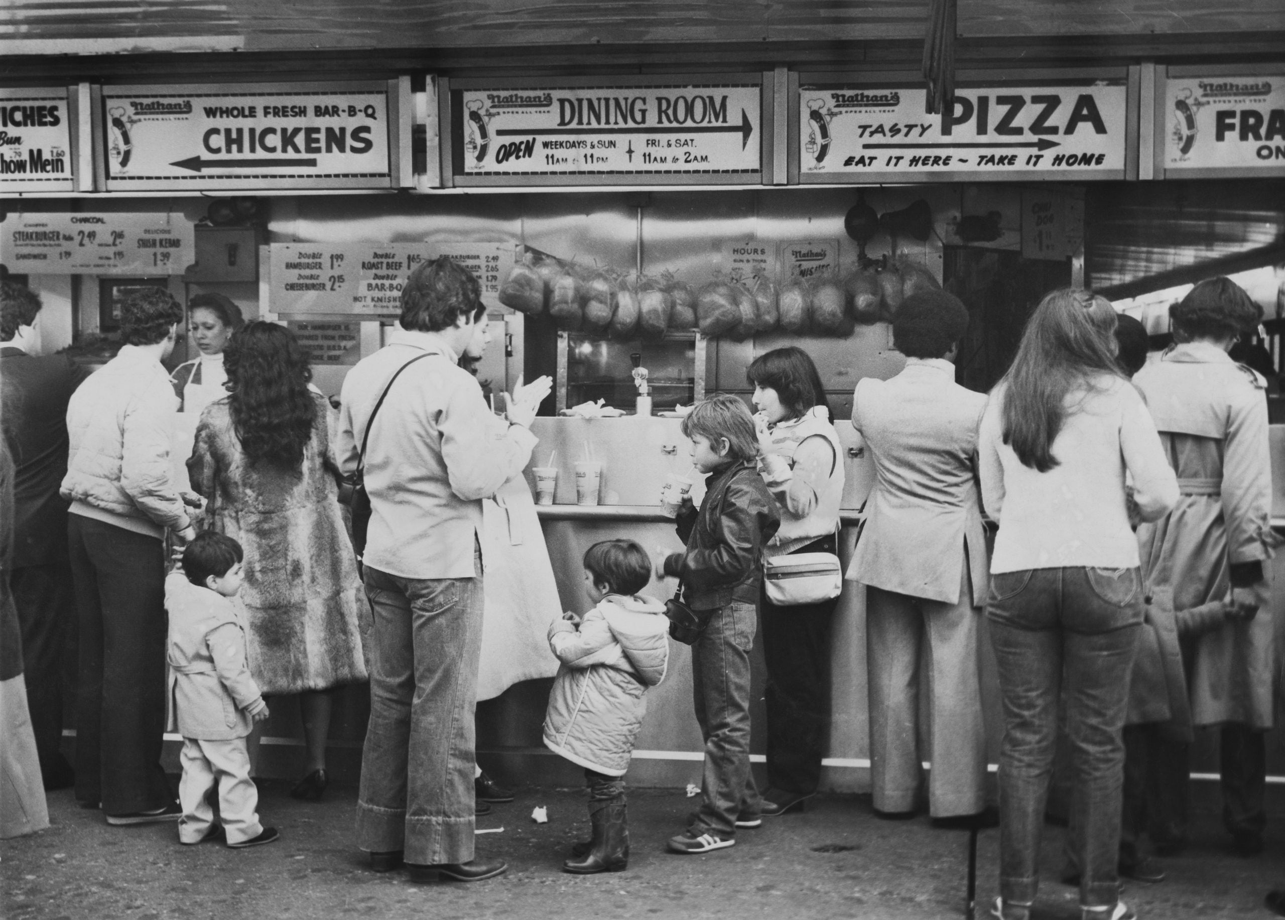 A line of people waiting to order food at a Nathan’s Famous restaurant counter with menu signs for barbecue chicken and pizza.