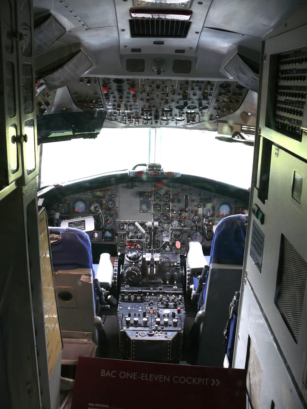 Cockpit of a BAC One-Eleven airplane showing numerous control panels, buttons, levers, and two blue pilot seats, viewed from the entrance with a placard that reads “BAC ONE-ELEVEN COCKPIT” in the foreground.