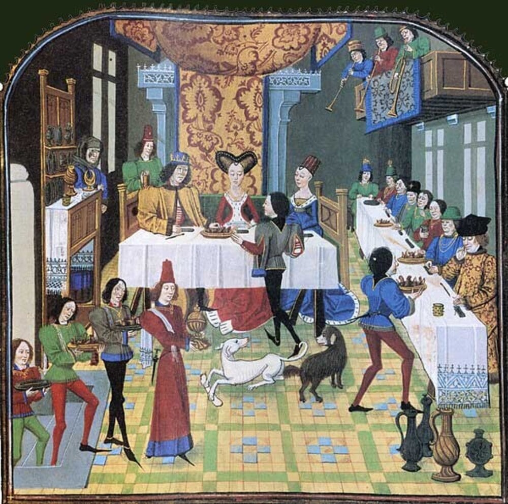Medieval banquet scene with royalty seated at a table, attendants serving food, musicians on a balcony, and two dogs on the floor.