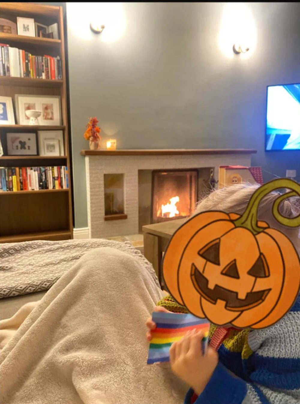 A person holding a rainbow-colored item with their face obscured by a digital pumpkin sticker, sitting on a couch in front of a lit fireplace and a tall bookshelf.