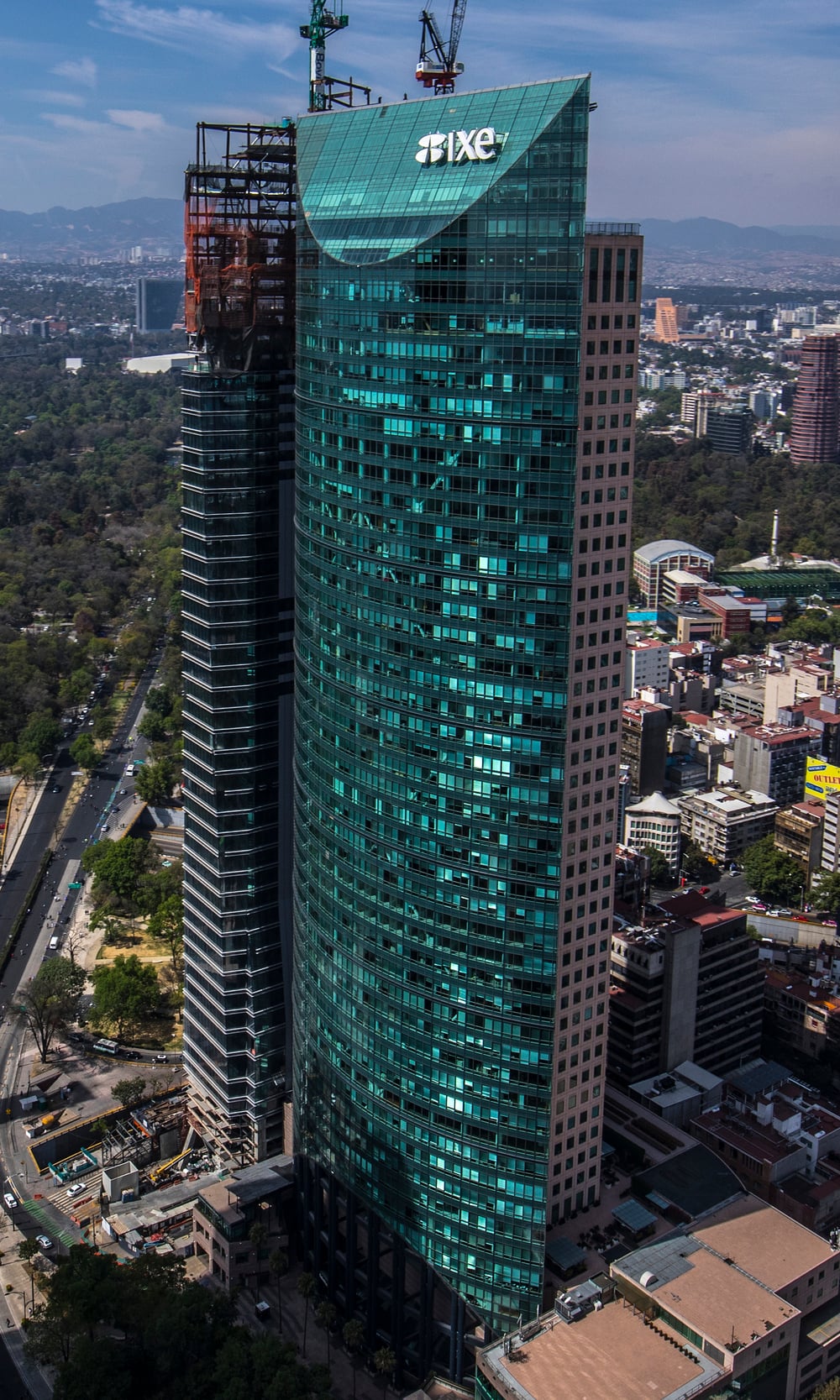 Aerial view of the Ixe skyscraper, a curved glass office building in Mexico City, with a construction crane visible atop an adjacent tower.