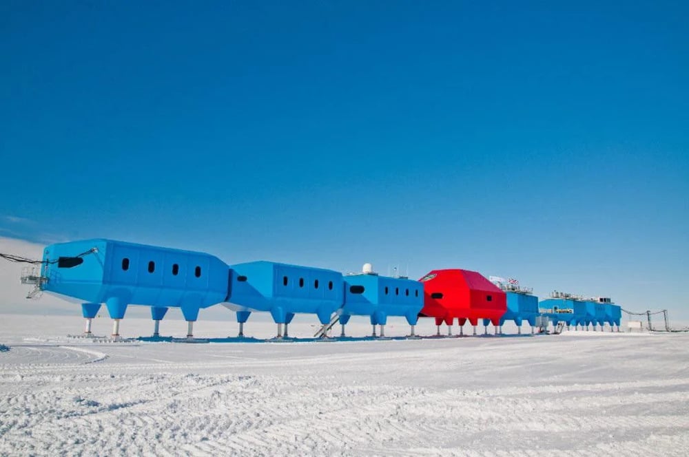 A line of interconnected, elevated blue and red research modules standing on stilts across a vast, snow-covered landscape under a clear blue sky.