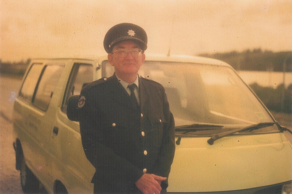 An older man in a dark police uniform and cap stands in front of a yellow van on a cloudy day. The photo has a faded, vintage look.