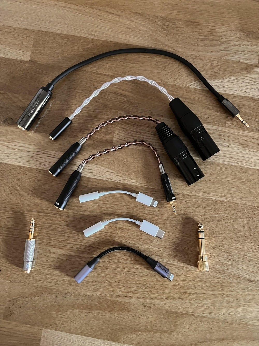 A variety of audio adapters and cables, including 3.5mm, XLR, Lightning, and USB-C connectors, are neatly arranged on a wooden surface.