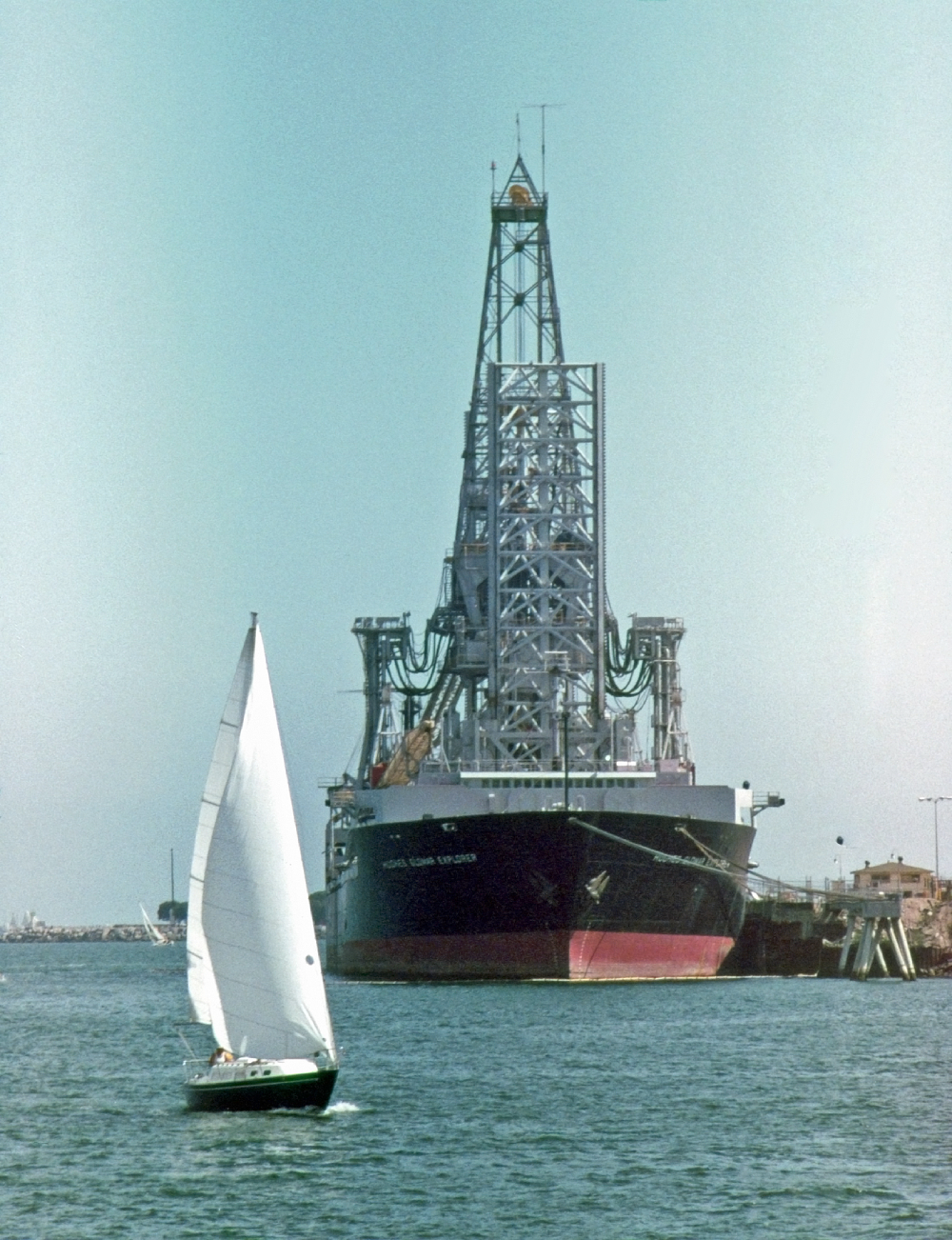 A small sailboat sails past the large drillship Hughes Glomar Explorer docked at a pier.