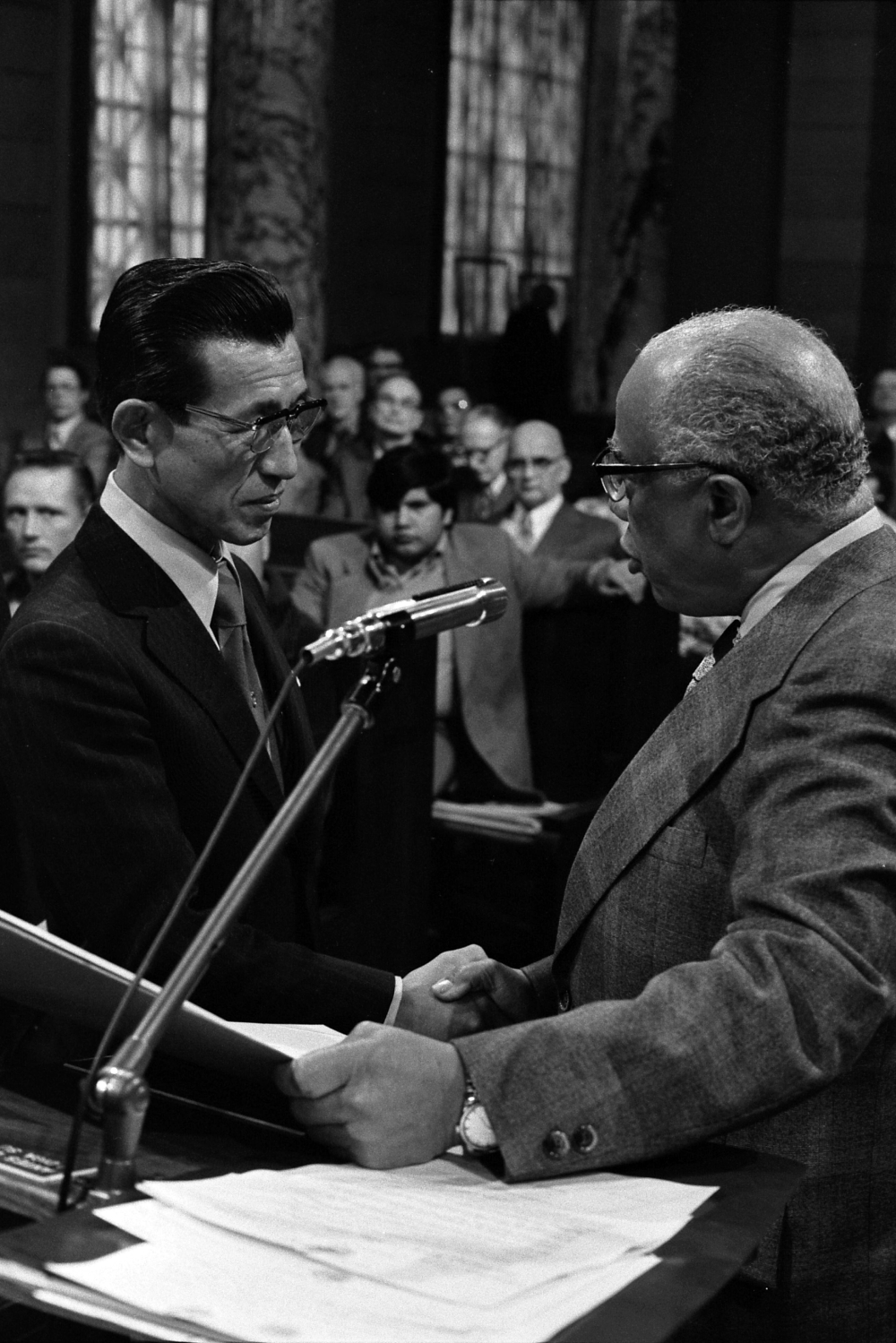 Two men in suits shake hands across a podium microphone during a hearing.