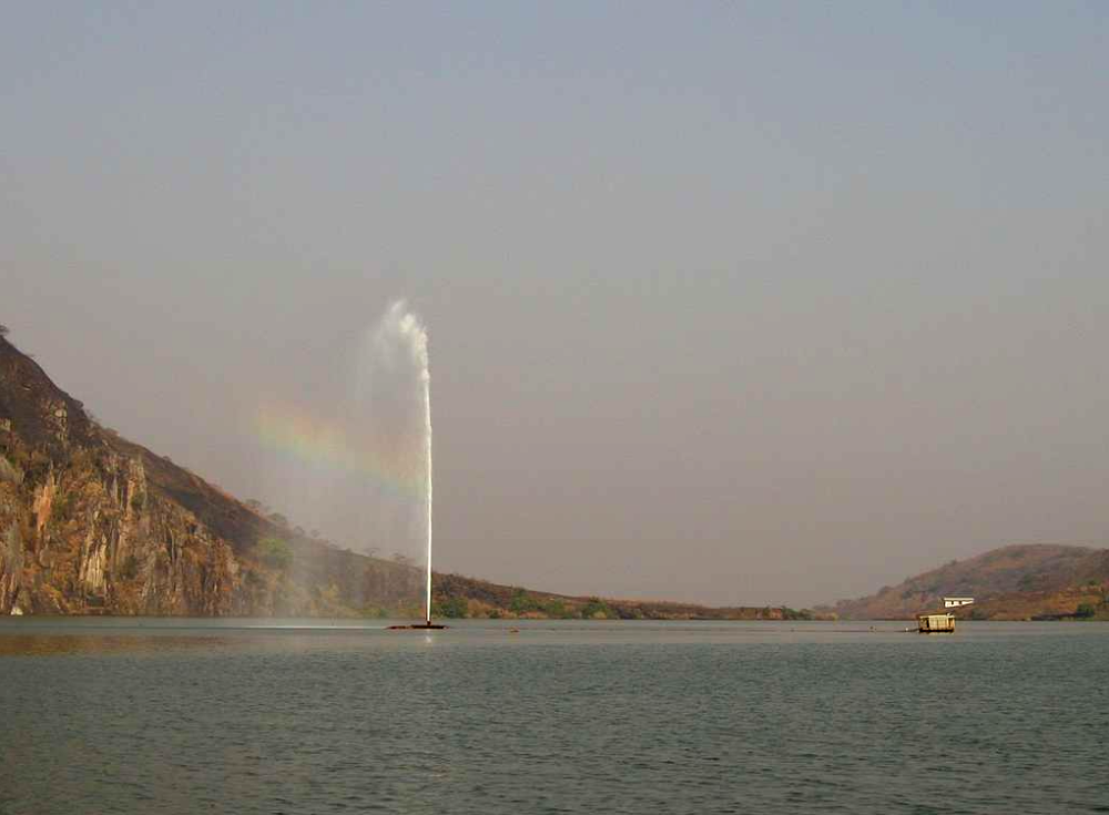 A fountain sprays a tall jet of water into a lake surrounded by hills, with a faint rainbow visible in the mist.