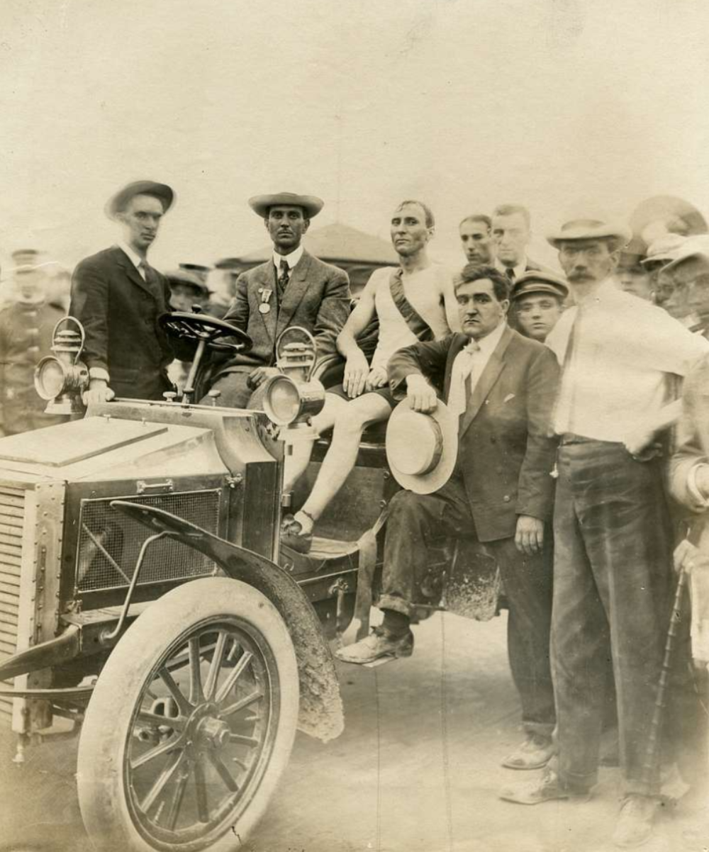 A group of men surrounding an early 20th-century automobile, with one man sitting in the driver’s seat wearing a tank top and a sash.