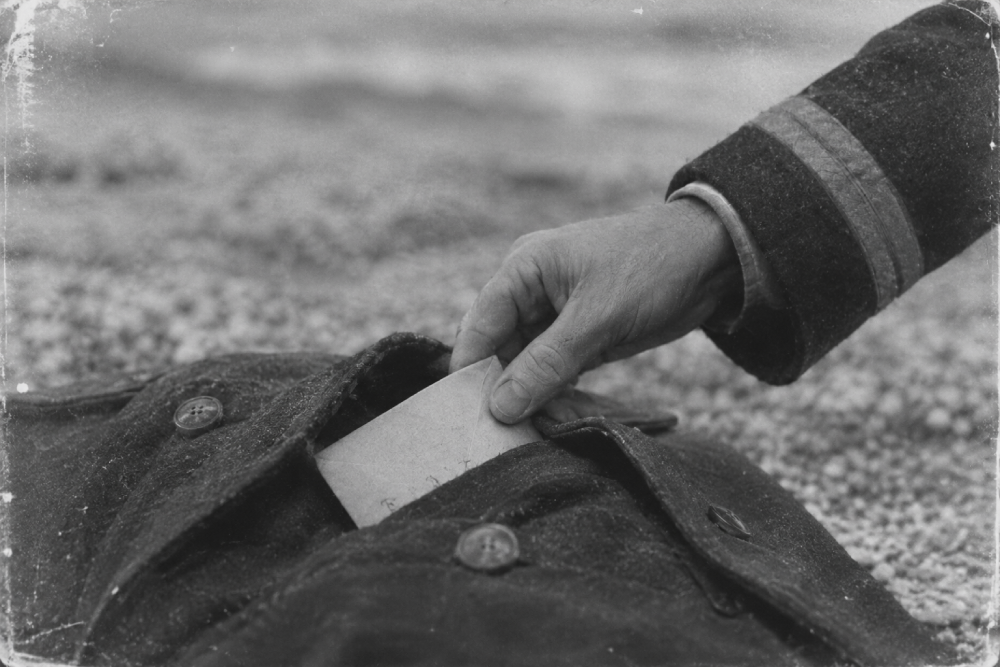 A person wearing a sleeve with stripes tucks an envelope into the pocket of a coat lying on a pebbled surface.