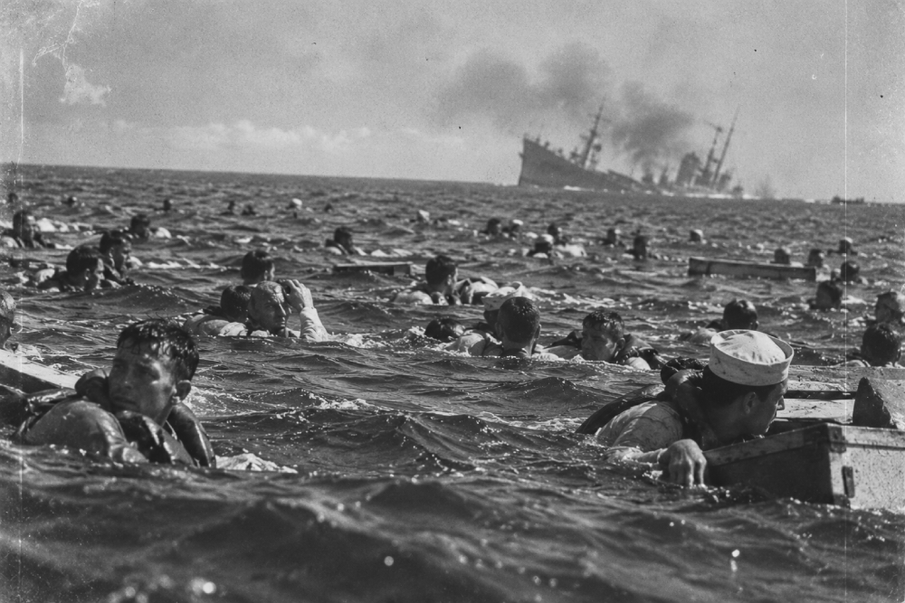 Many sailors in the water, some wearing life vests and holding onto wooden crates, with a smoking ship sinking in the distance.