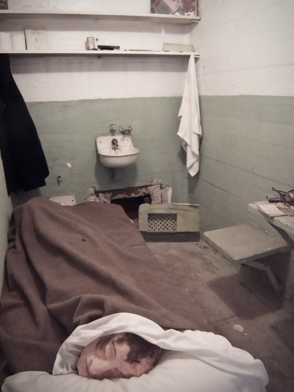 A mannequin head rests on a pillow under a brown blanket in a sparse, green-walled prison cell with a small sink.