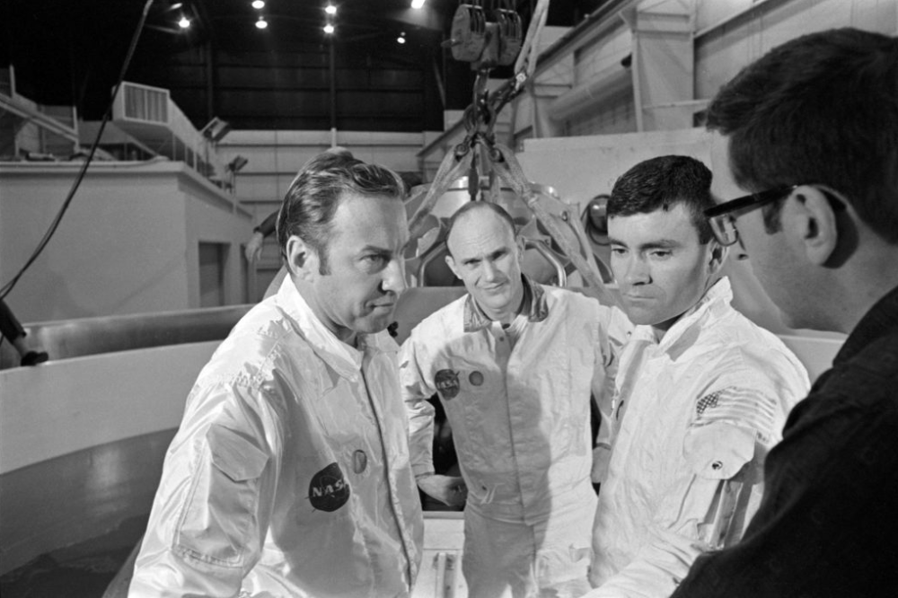 Three NASA astronauts in white flight suits stand in a facility, appearing to listen to a man in the foreground.