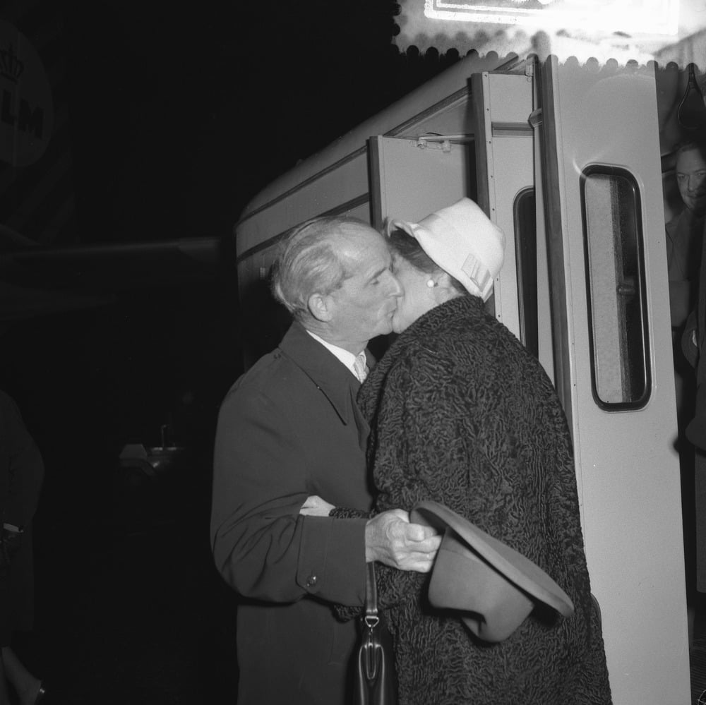 An older man and woman share a kiss goodbye outside a bus at night. The man holds a hat and the woman wears a textured coat and a white hat. People and part of a bus are visible in the background.