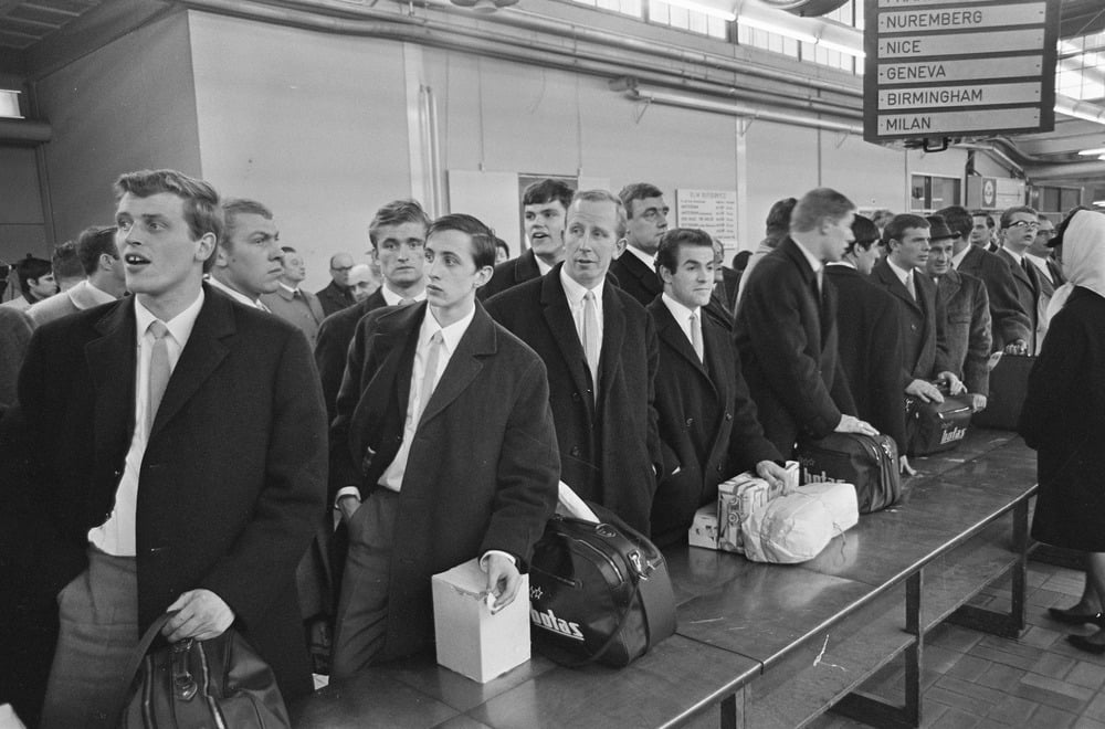 A group of young men in coats stand in line at an airport check-in counter, carrying bags and boxes. Overhead signs list European cities like Nice, Geneva, and Milan. The mood appears orderly and anticipatory.