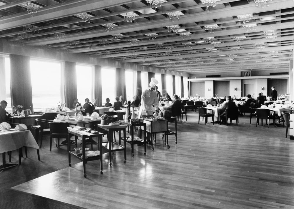 Black and white photo of a spacious restaurant with large windows, diners seated at tables, and waitstaff serving food. The room has a patterned ceiling and wooden floor, creating a formal, mid-20th-century atmosphere.