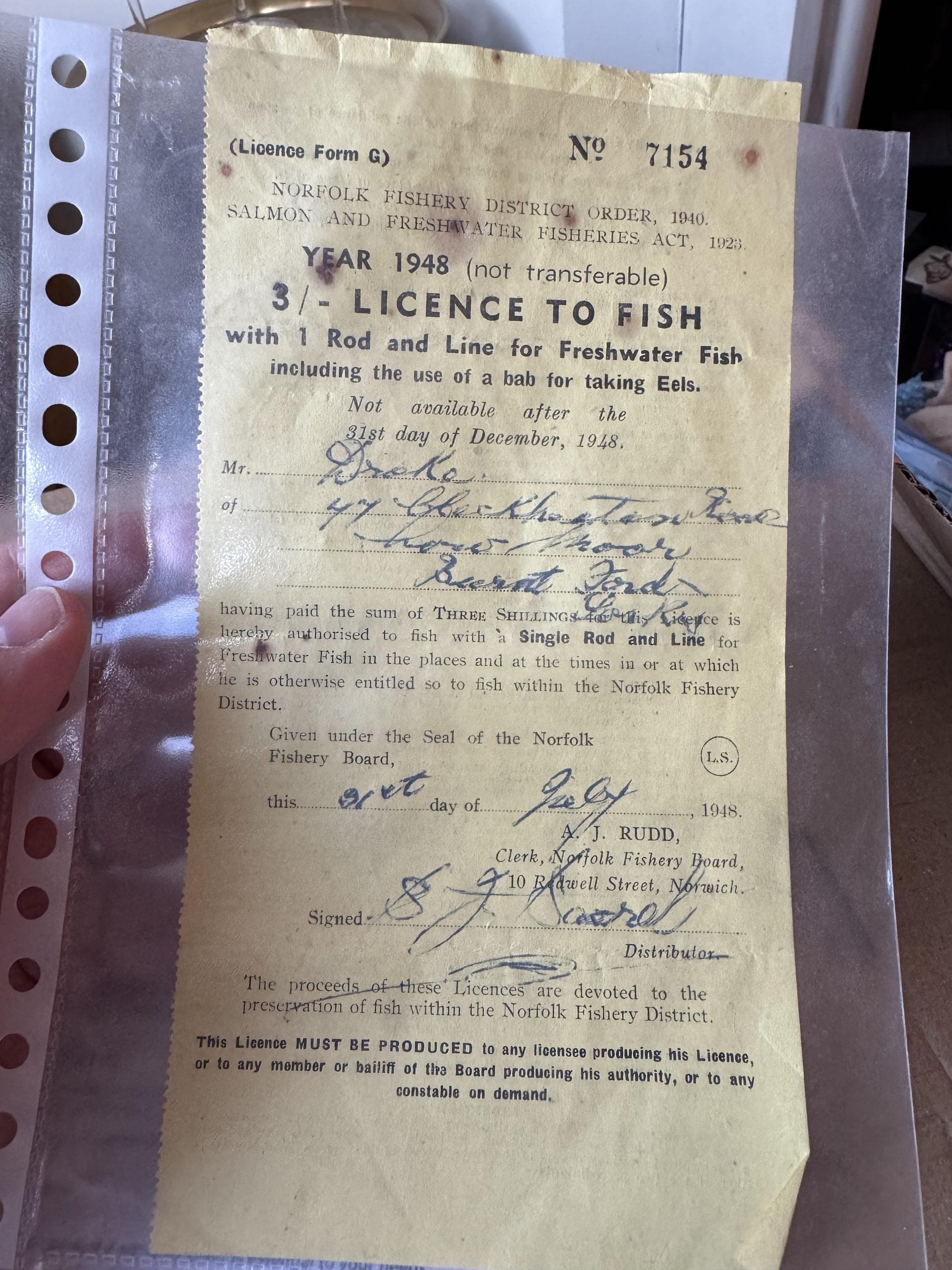 A 1948 Norfolk Fishery District fishing licence form, numbered 7154, printed on yellow paper and protected in a plastic sleeve.