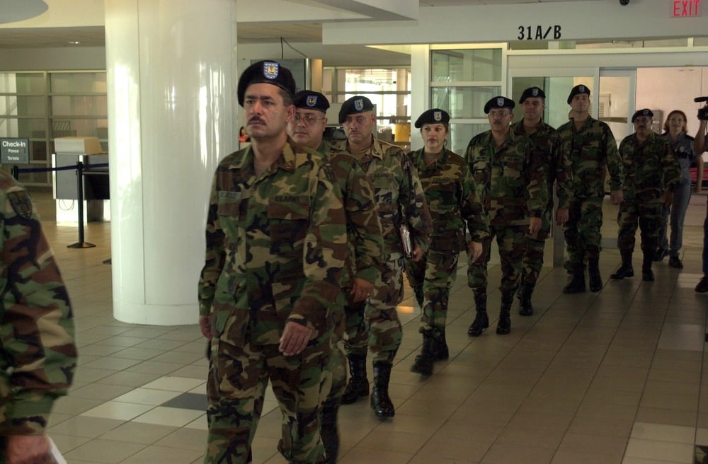 A group of uniformed soldiers in camouflage walk in formation through an indoor public space with tiled floors and white columns.