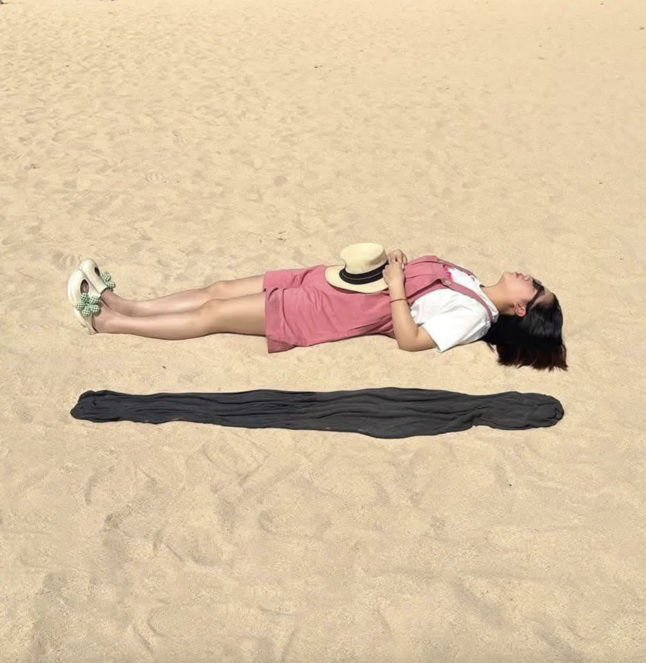 A person lying on their back on the sand, with a long, dark fabric strip placed parallel to their body.