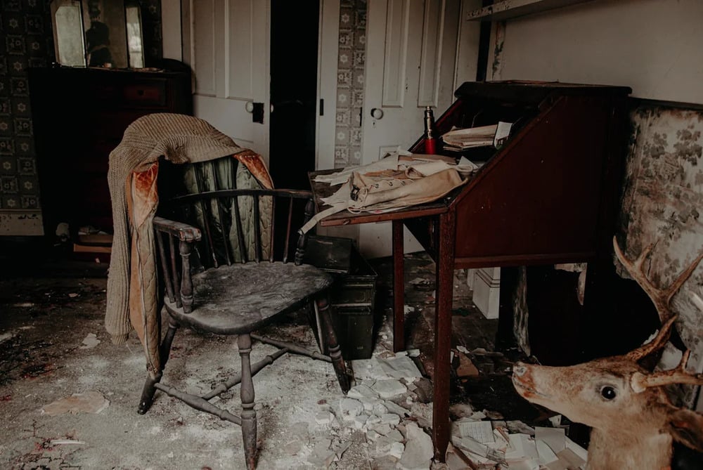 A wooden chair with a draped sweater sits next to an antique writing desk in a room with debris on the floor and a deer mount in the foreground.