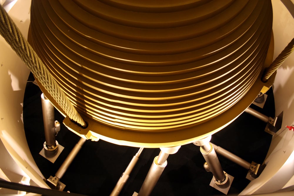 A large, ribbed, golden-colored tuned mass damper suspended by thick cables above support struts.