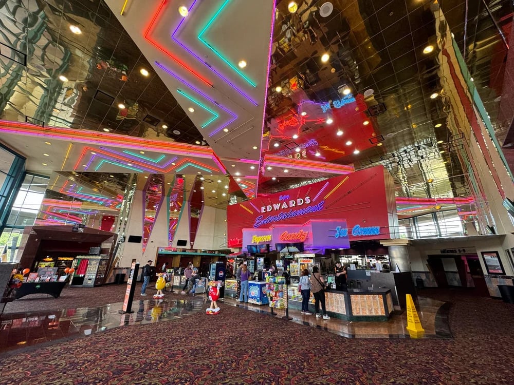 The brightly lit interior of a movie theater lobby with a neon-accented concession stand under a reflective mirrored ceiling.