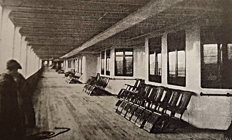 A long wooden deck on a ship with rows of empty chairs lined up against a wall of windows and a person standing in the foreground.