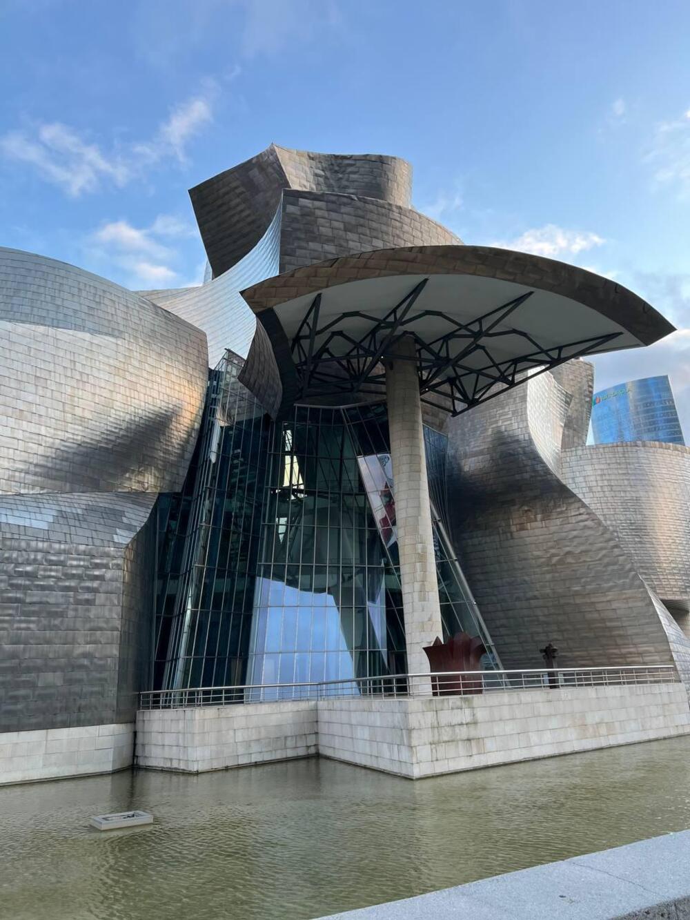 The Guggenheim Museum Bilbao, featuring its signature titanium-clad curves and glass atrium, viewed from the water’s edge.