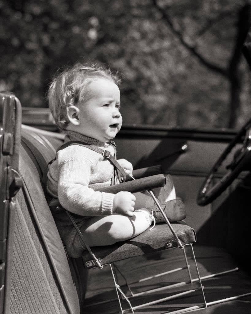 A young child in a sweater sits in a metal-framed car seat mounted on the front passenger seat of a vintage vehicle.