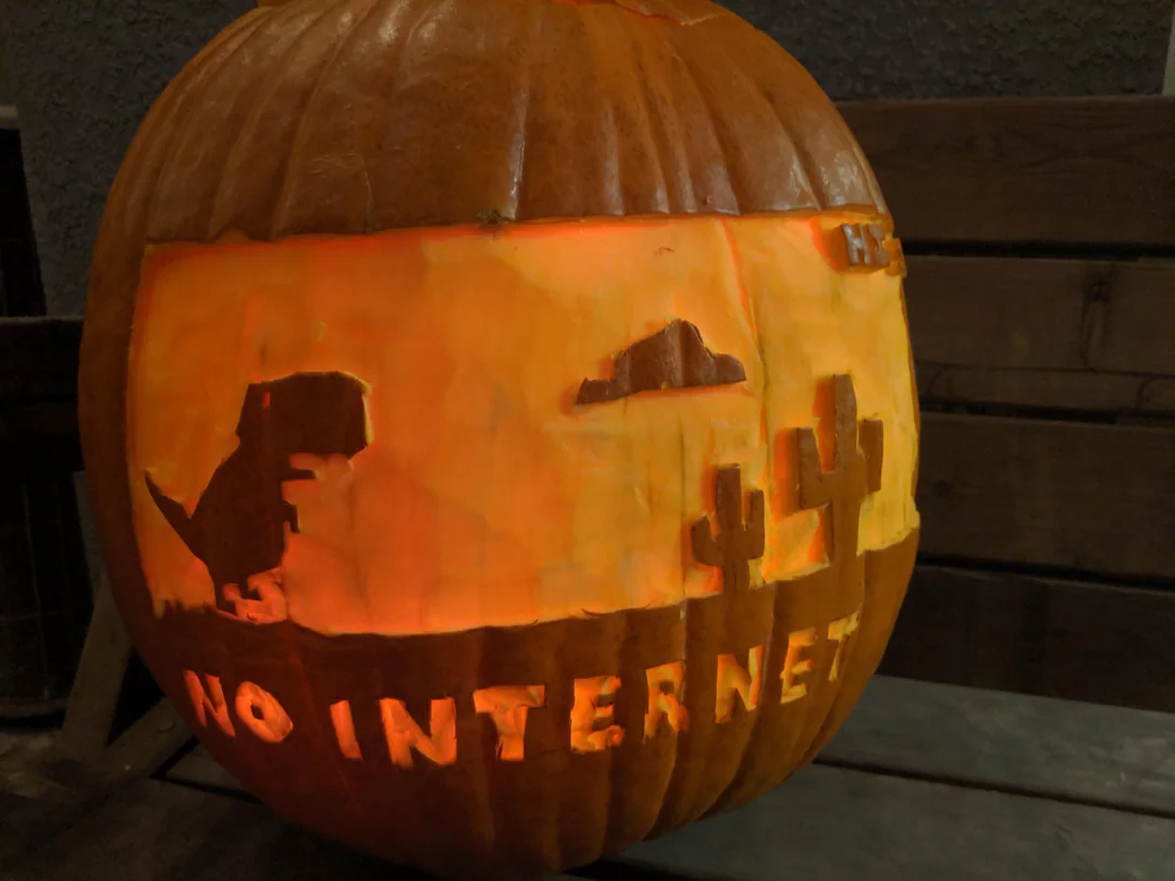 A carved pumpkin lit from within, featuring the silhouette of the Chrome dinosaur game, a cactus, and the text “NO INTERNET.”