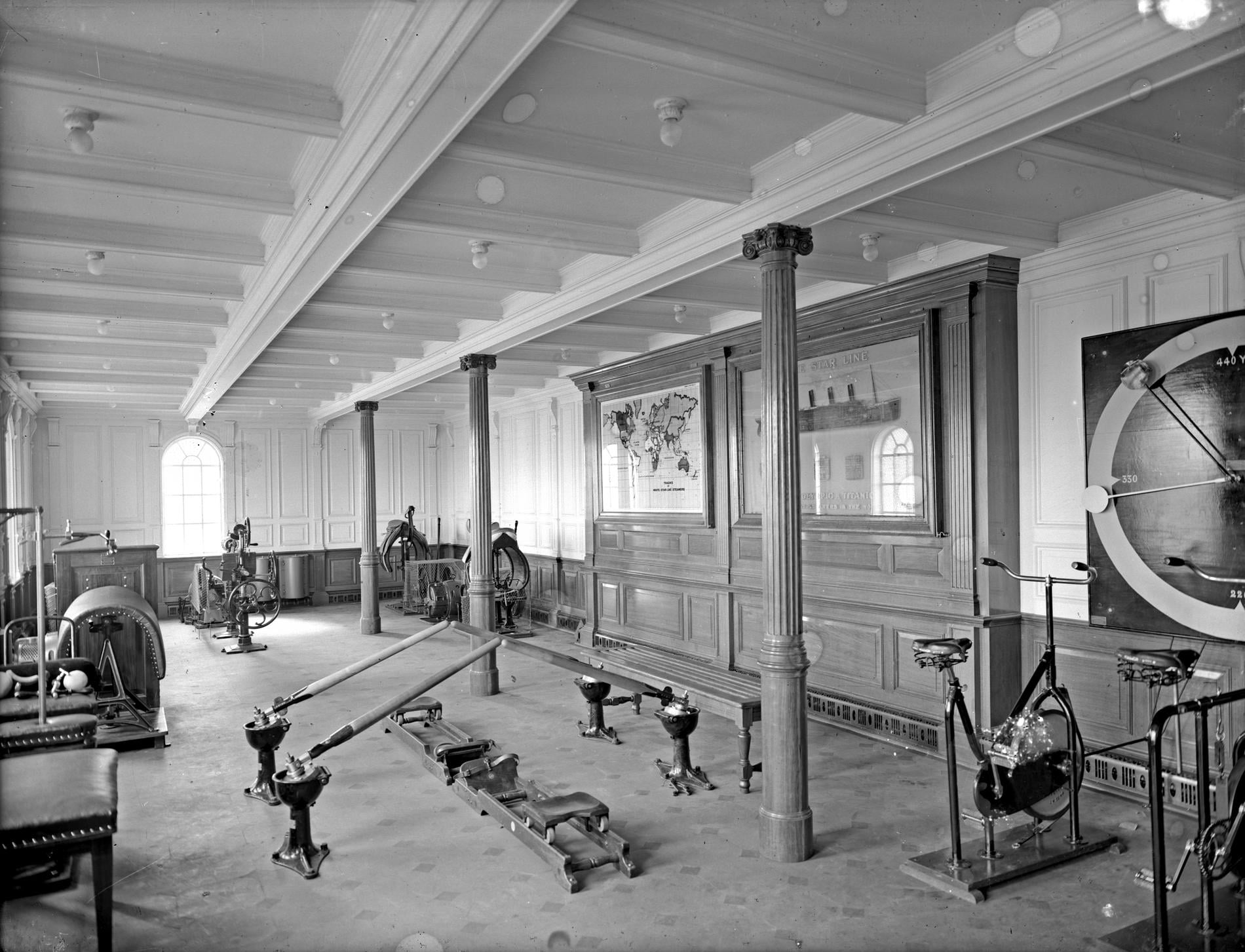 The gymnasium on the RMS Titanic, featuring rowing machines, stationary exercise bicycles, and a wall-mounted map and White Star Line poster.