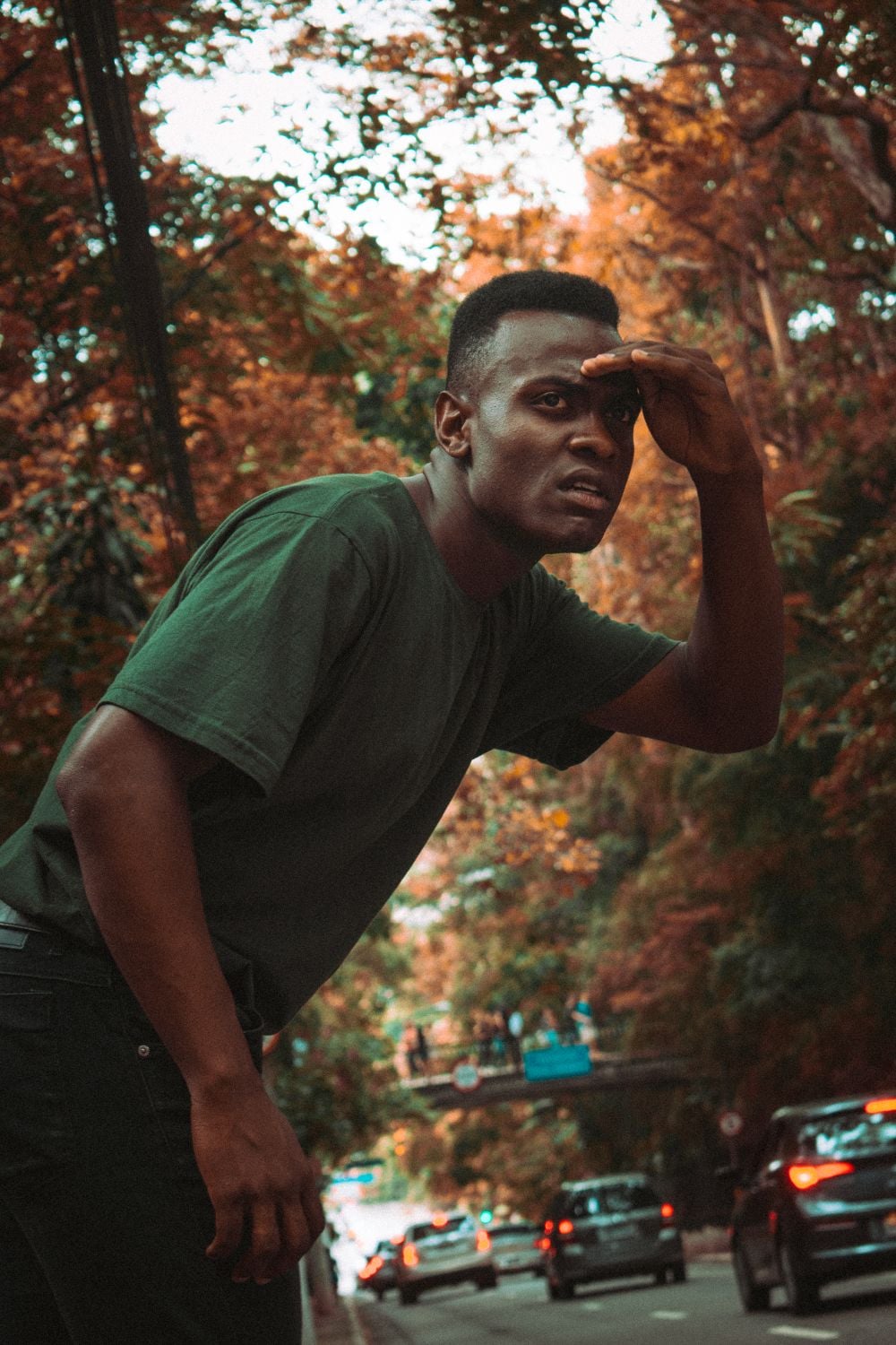 A man in a dark shirt leans forward and shades his eyes with his hand while standing on a tree-lined street with cars in the background. Autumn leaves color the scene.