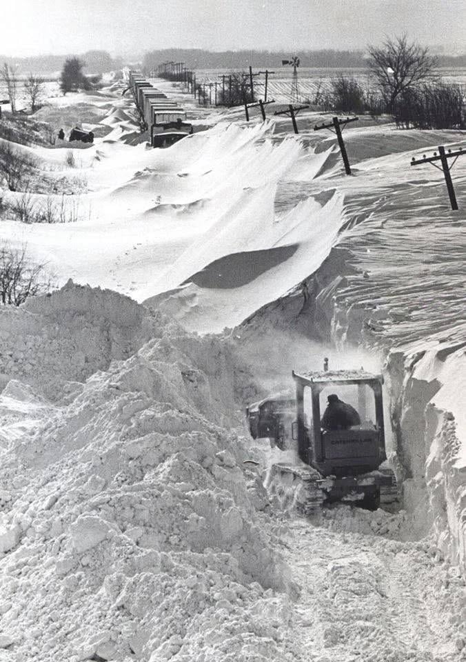A bulldozer clears a deep, wind-swept snowdrift from a rural road, with a line of train cars visible in the distance.