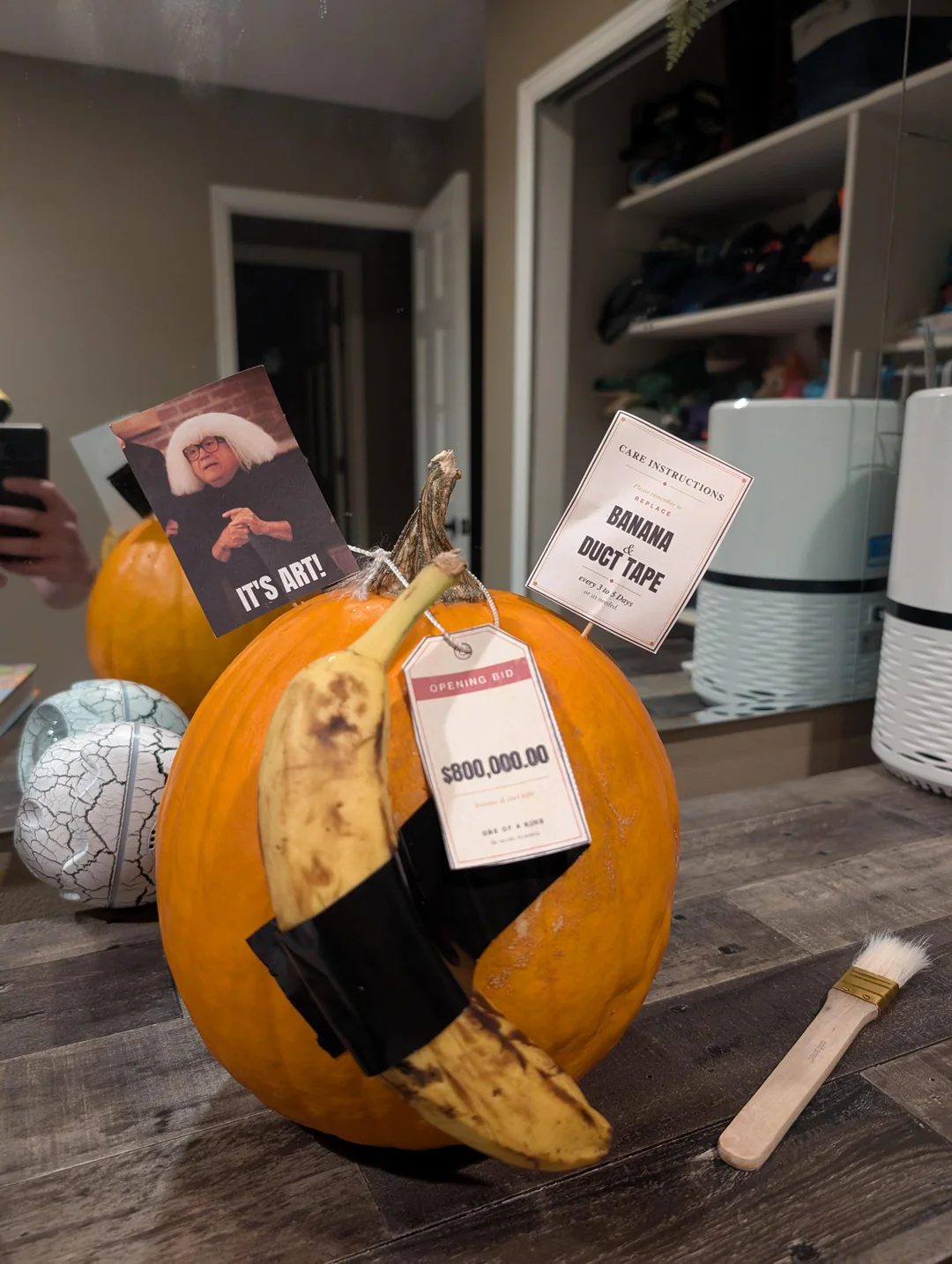 A pumpkin with a banana taped to it, adorned with a picture of Danny DeVito, an opening bid tag for $800,000.00, and a care instructions tag for “Banana & Duct Tape.”