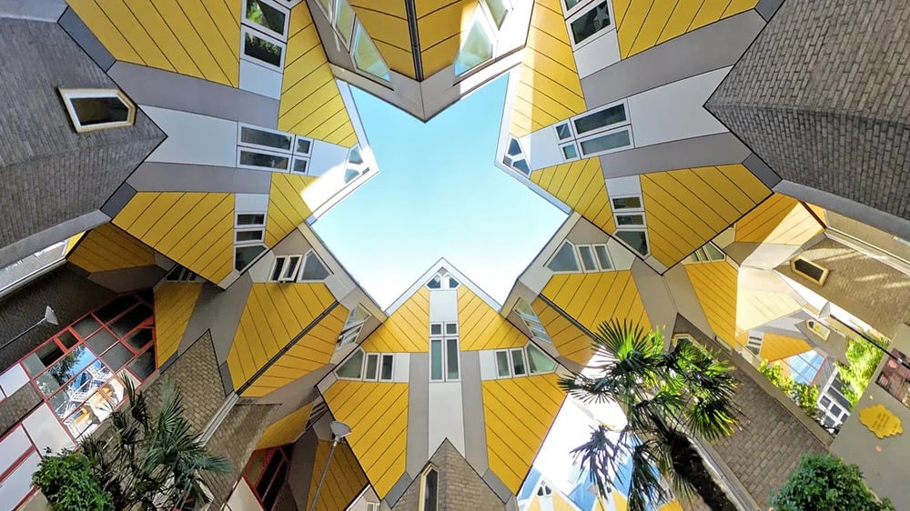 Looking upward at the Cube Houses in Rotterdam, featuring geometric yellow buildings angled toward a star-shaped opening to the sky.