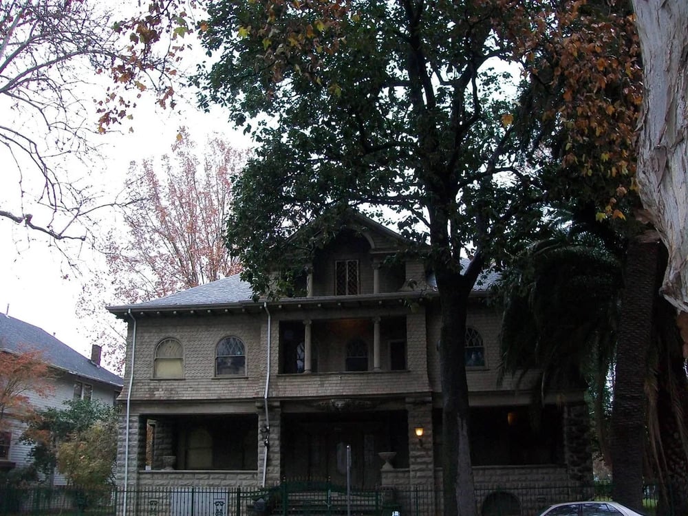 A two-story stone house with a covered front porch and arched windows, framed by mature trees.