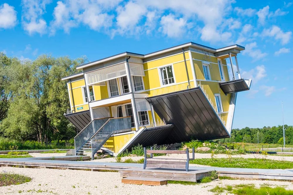 A yellow two-story house sitting upside down on its roof in a park.
