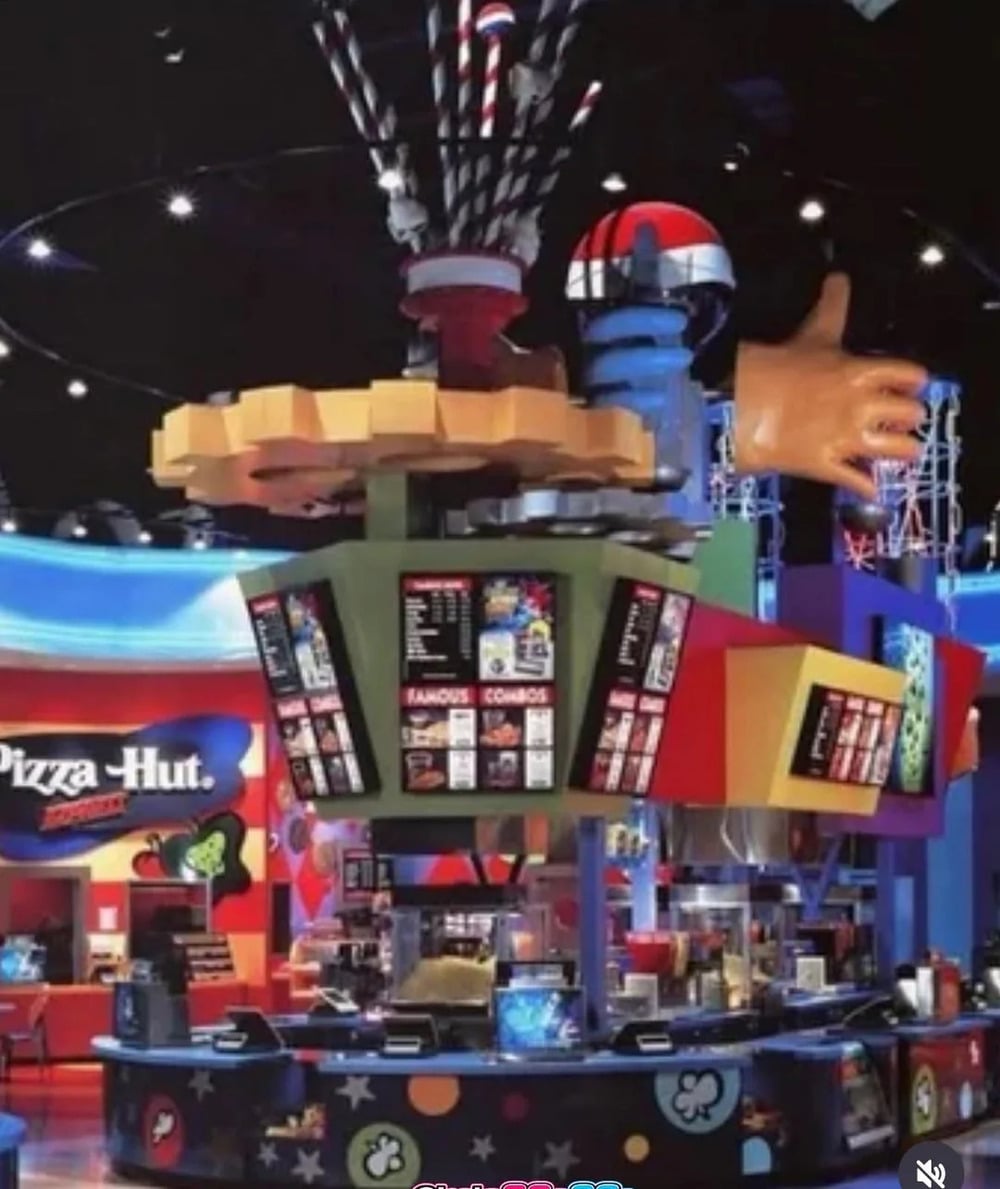 A brightly colored Pizza Hut Express food court kiosk with a central overhead menu display and a large sculptural hand reaching out.