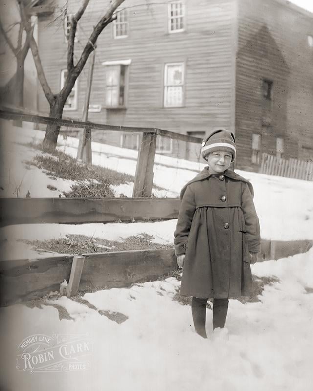 A young child wearing a winter coat and hat stands in the snow in front of a wooden fence and a house.