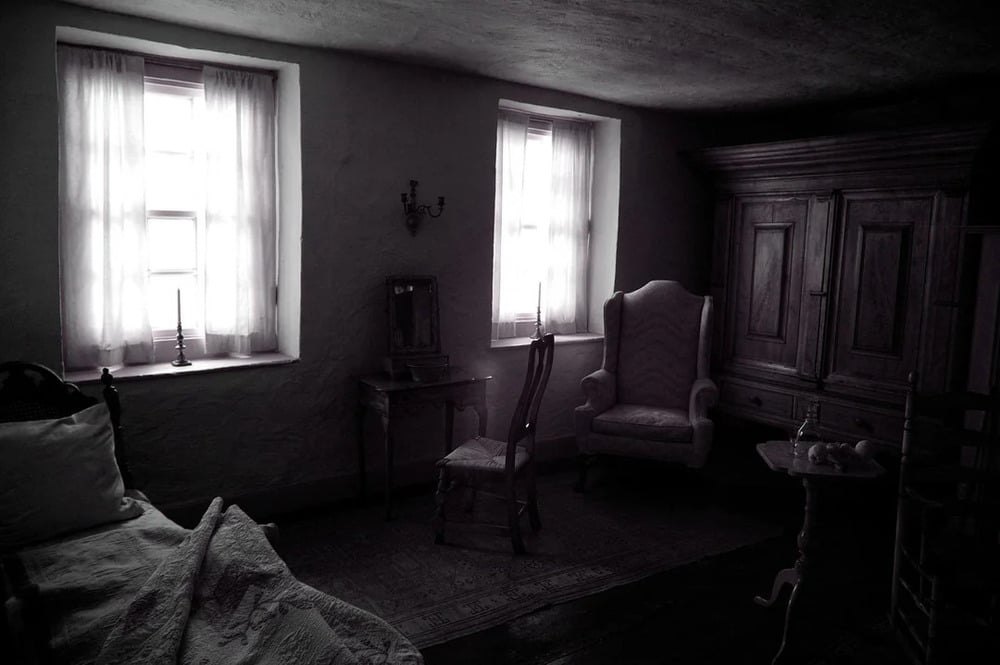 A dimly lit room featuring a bed with a quilt, a vanity table with a chair, a high-backed armchair, and a large wooden cabinet, all illuminated by two windows with white curtains.