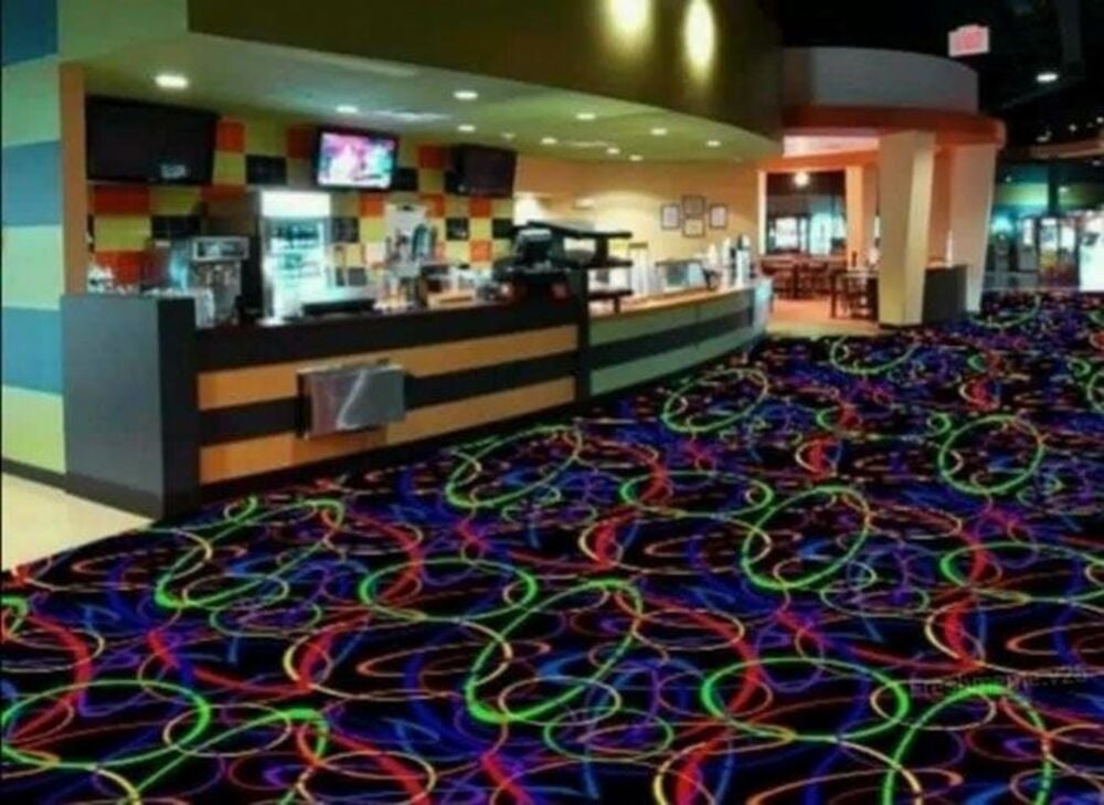 A concession stand with a service counter and overhead televisions, set against a floor featuring a black carpet with a neon-colored swirl pattern.