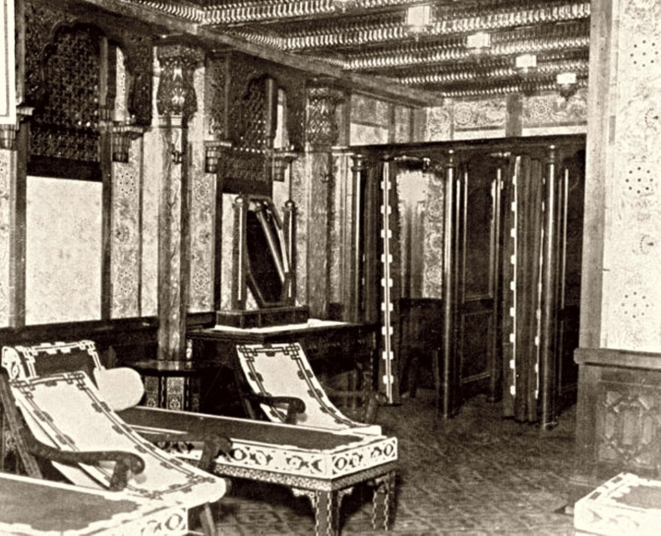 Vintage photograph of an ornate, wood-paneled room on the Titanic featuring patterned wallpaper, two lounge chairs, and a series of curtained changing stalls.