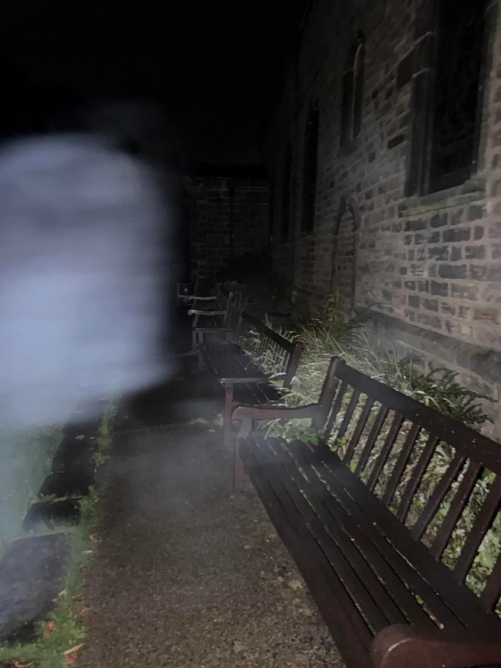 A row of wooden benches sits along a stone building at night, with a blurry, translucent shape in the foreground.