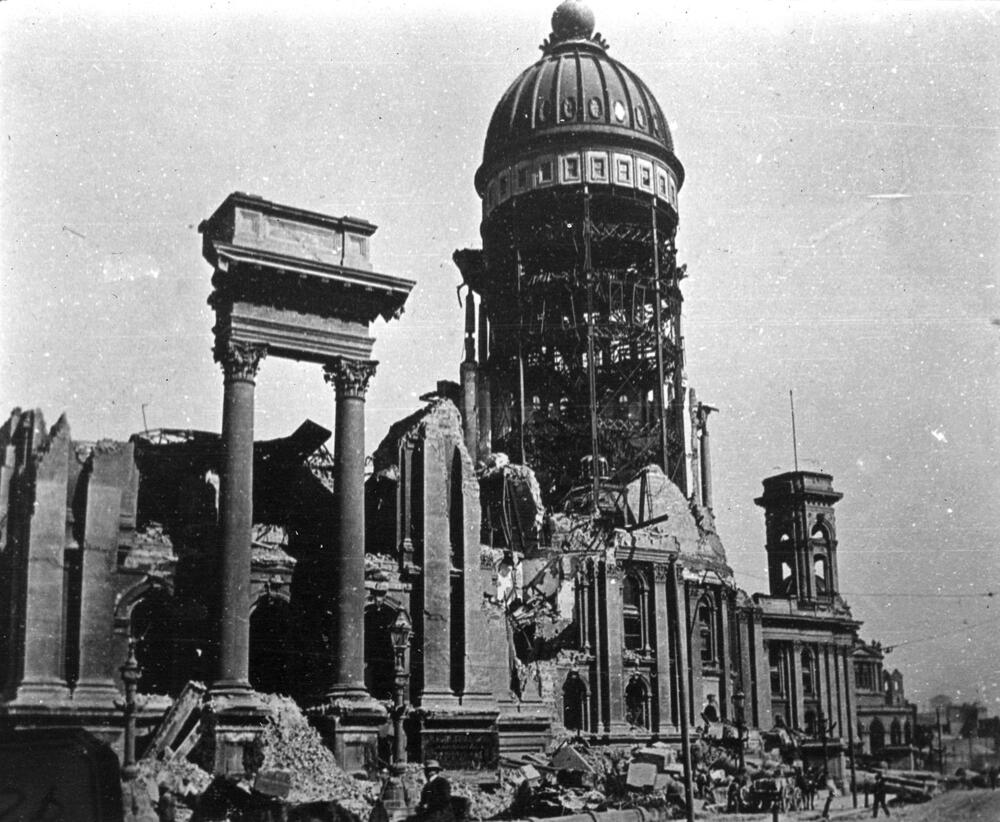 A black and white photograph of a severely damaged, domed public building with crumbled walls and exposed interior scaffolding following a disaster.