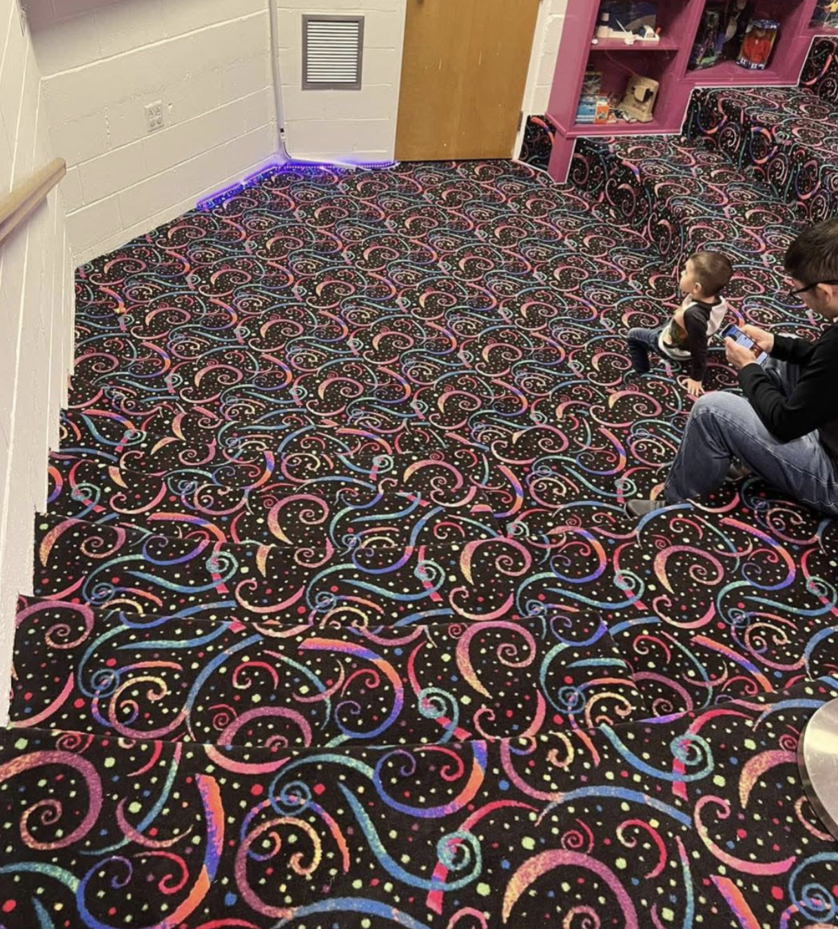 An adult and a young child sit on steps covered in black carpet with a bright, multicolored swirl and dot pattern.
