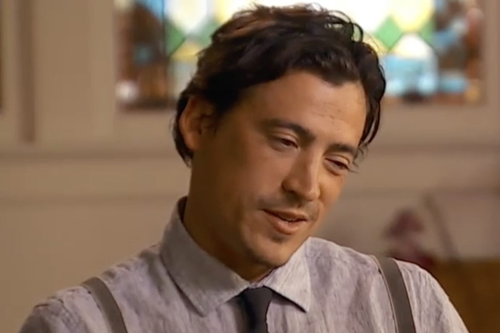 A man with dark hair, wearing a button-down shirt and suspenders, looking downward with a pensive expression.