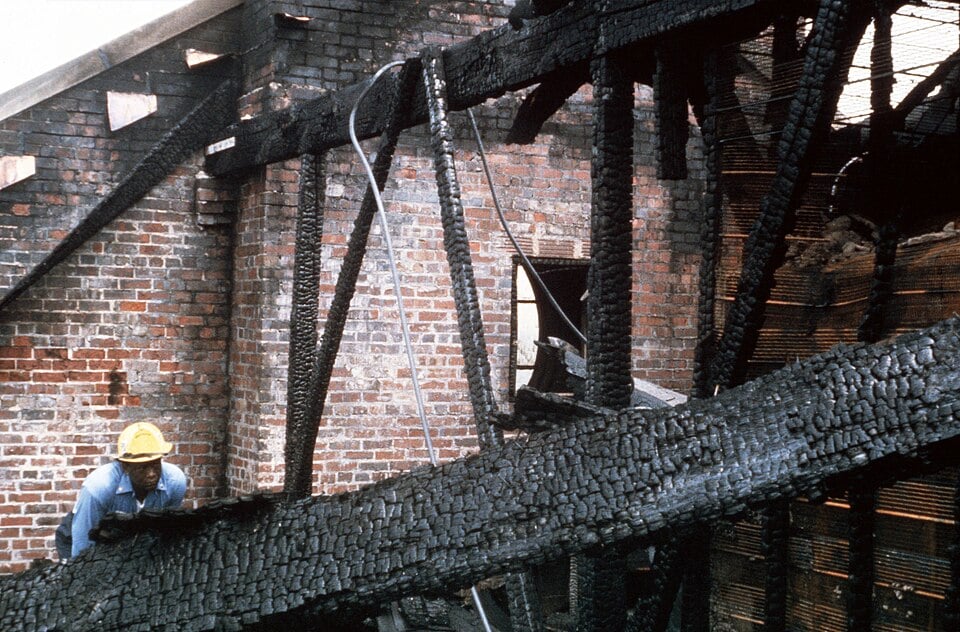 A worker in a yellow hard hat examines the charred wooden beams of a fire-damaged brick building.