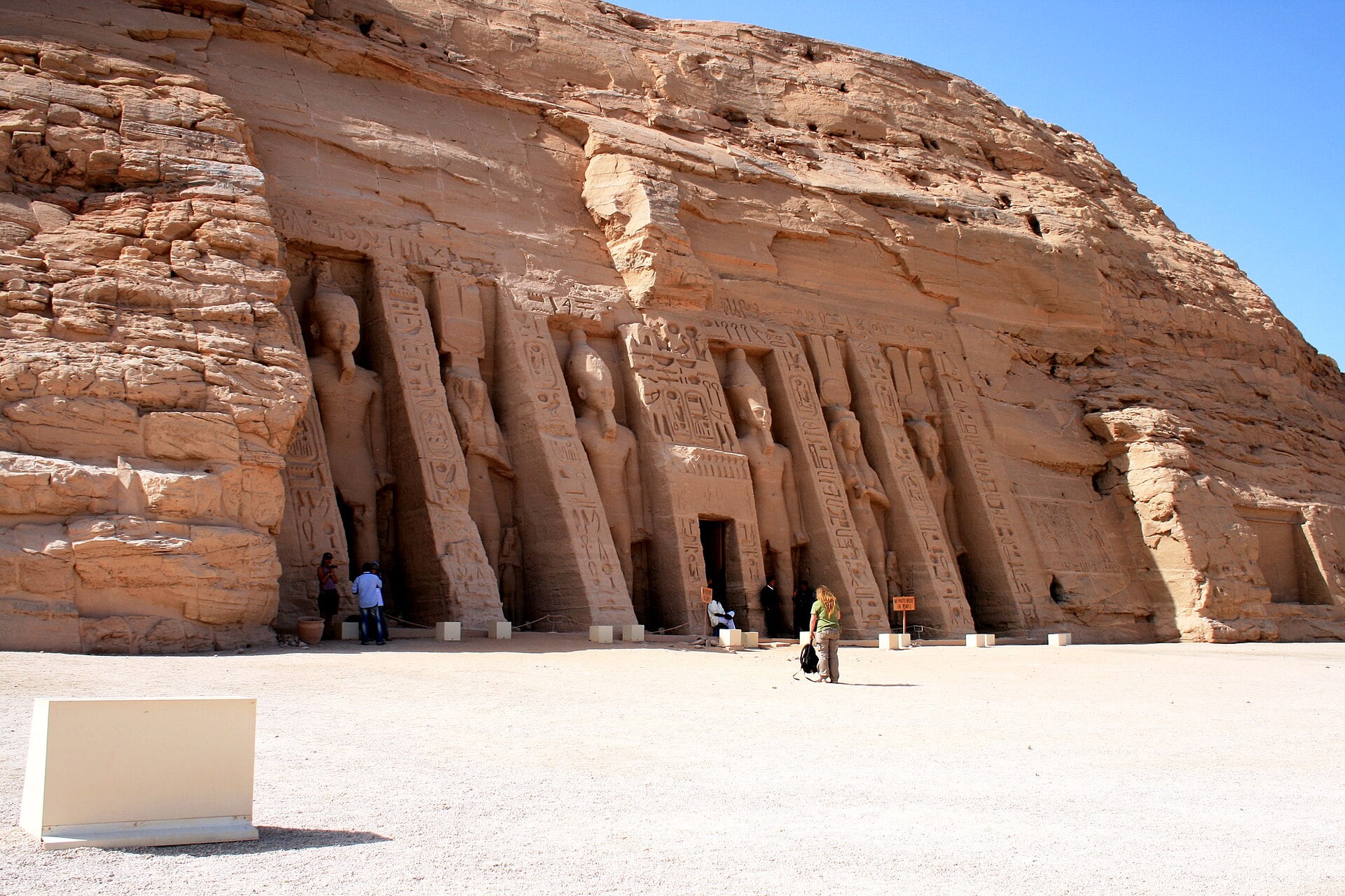 Large rock-cut statues of Ramses II carved into the cliff face of the Abu Simbel temples in Egypt, with tourists walking near the entrance.
