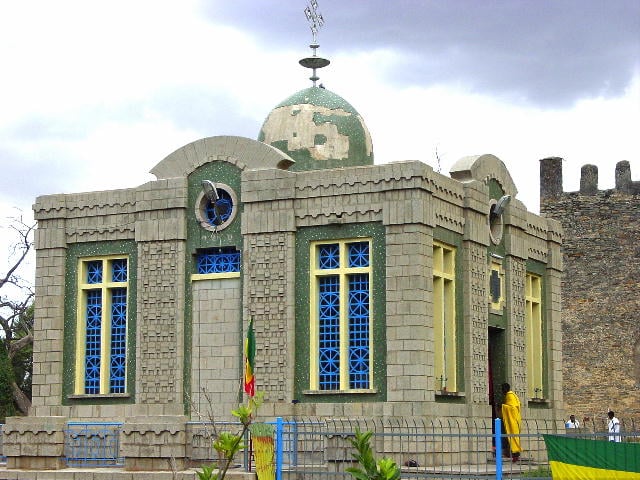 Stone chapel with arched blue windows and a dome, next to a stone wall.