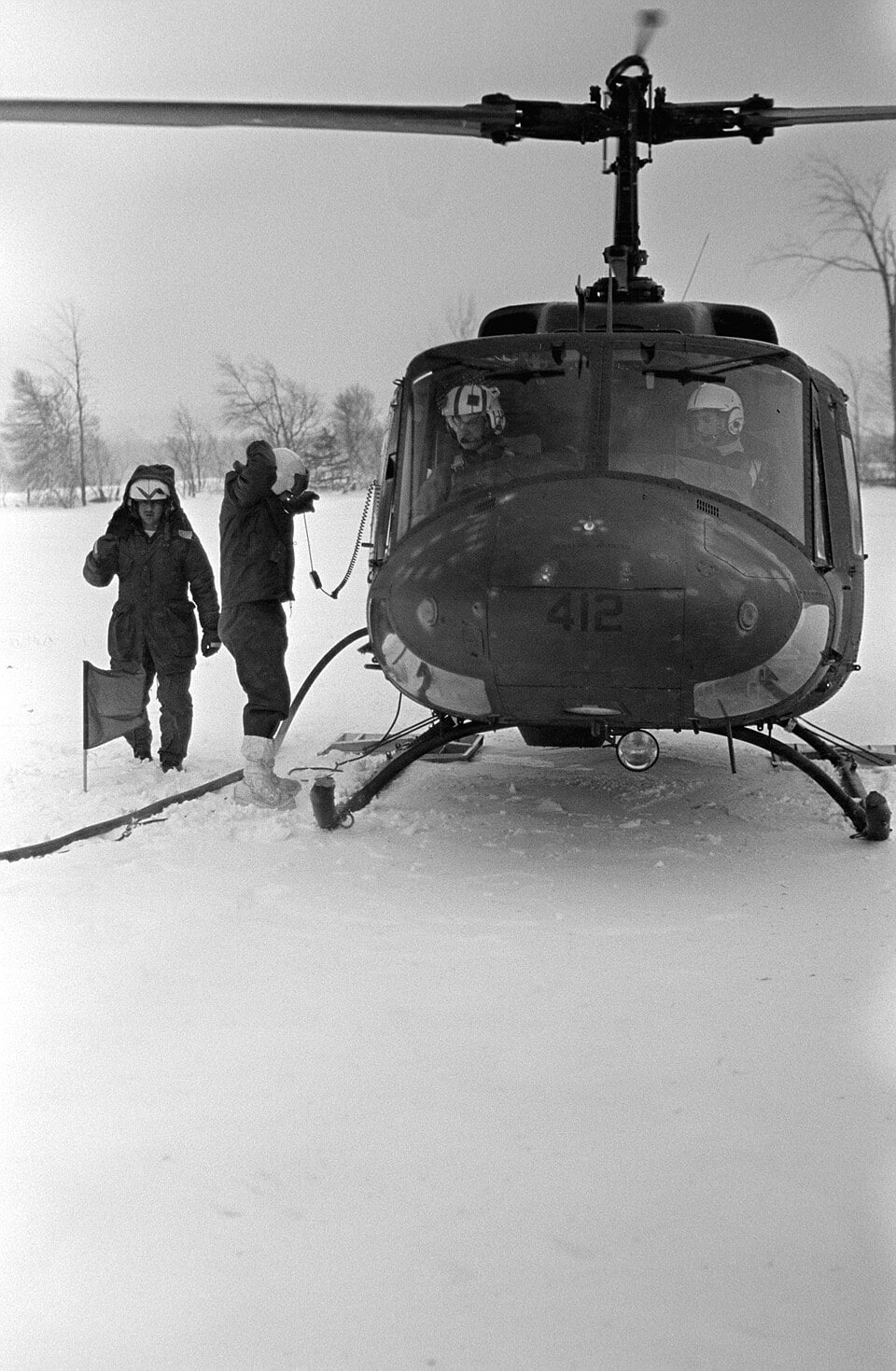 Two soldiers in winter gear stand in the snow next to a military helicopter marked with the number 412.