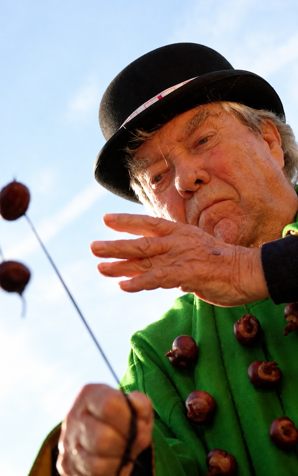 An older man in a black bowler hat and green coat decorated with conkers, holding a string with conkers attached.